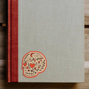 A hardcover book or journal with a textured light gray cover and a red cloth spine, placed on a wooden surface. A decorative sugar skull illustration appears near the lower left of the cover
