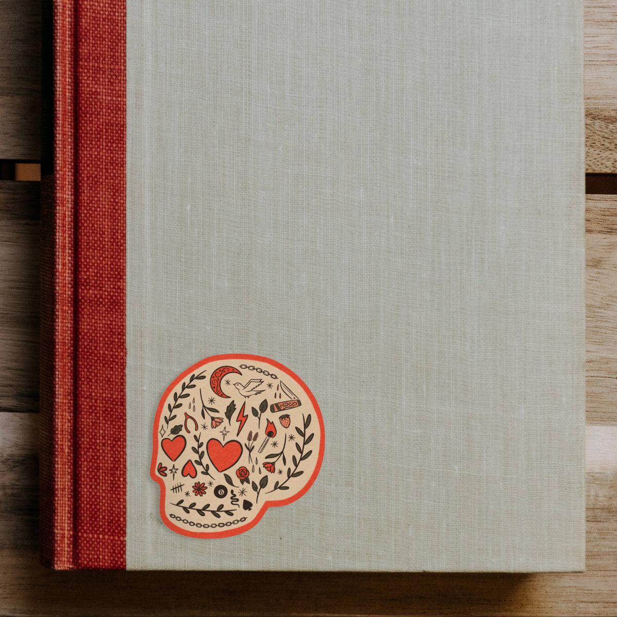A hardcover book or journal with a textured light gray cover and a red cloth spine, placed on a wooden surface. A decorative sugar skull illustration appears near the lower left of the cover