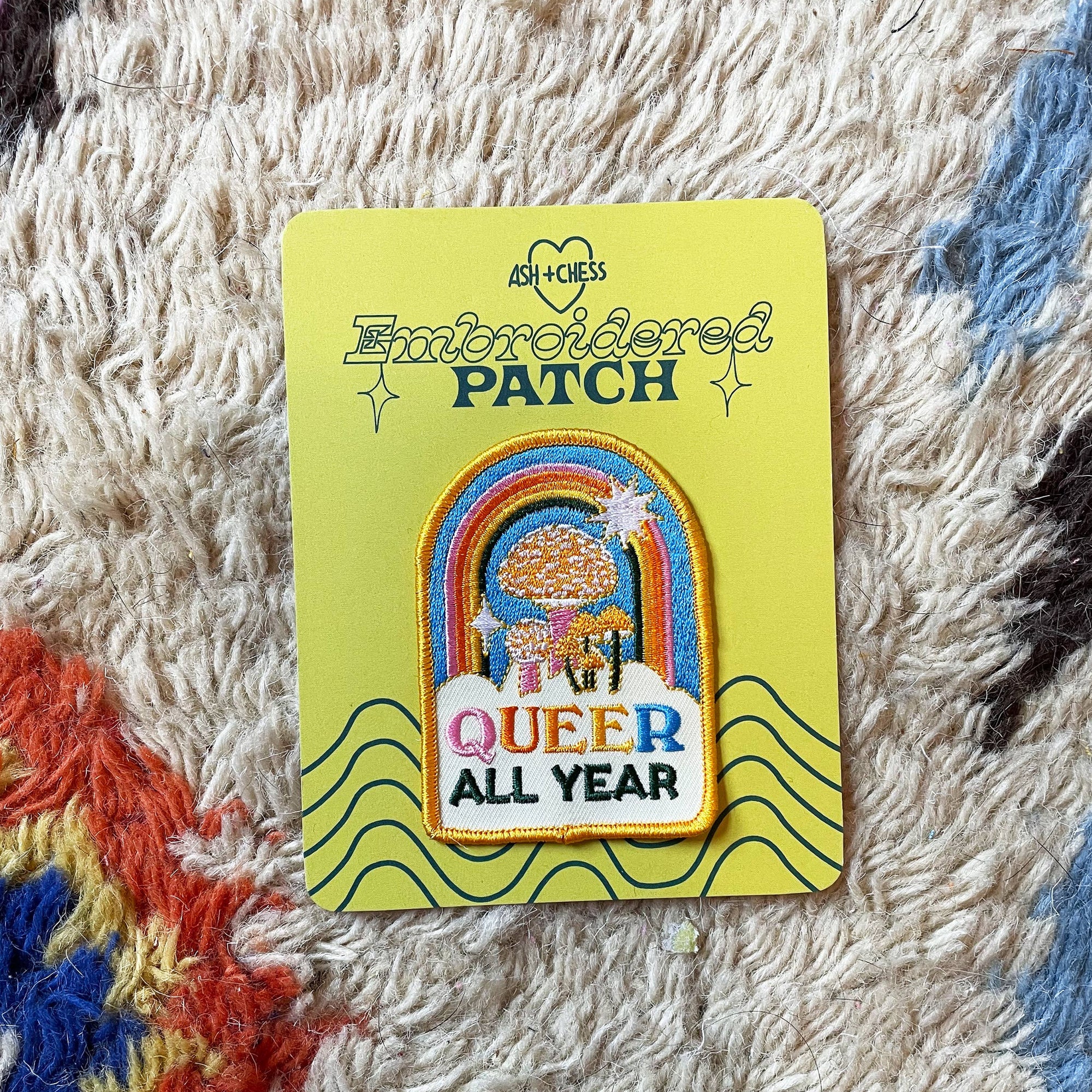 Embroidered patch on a yellow card with 'Queer All Year' text, displayed on a textured surface.