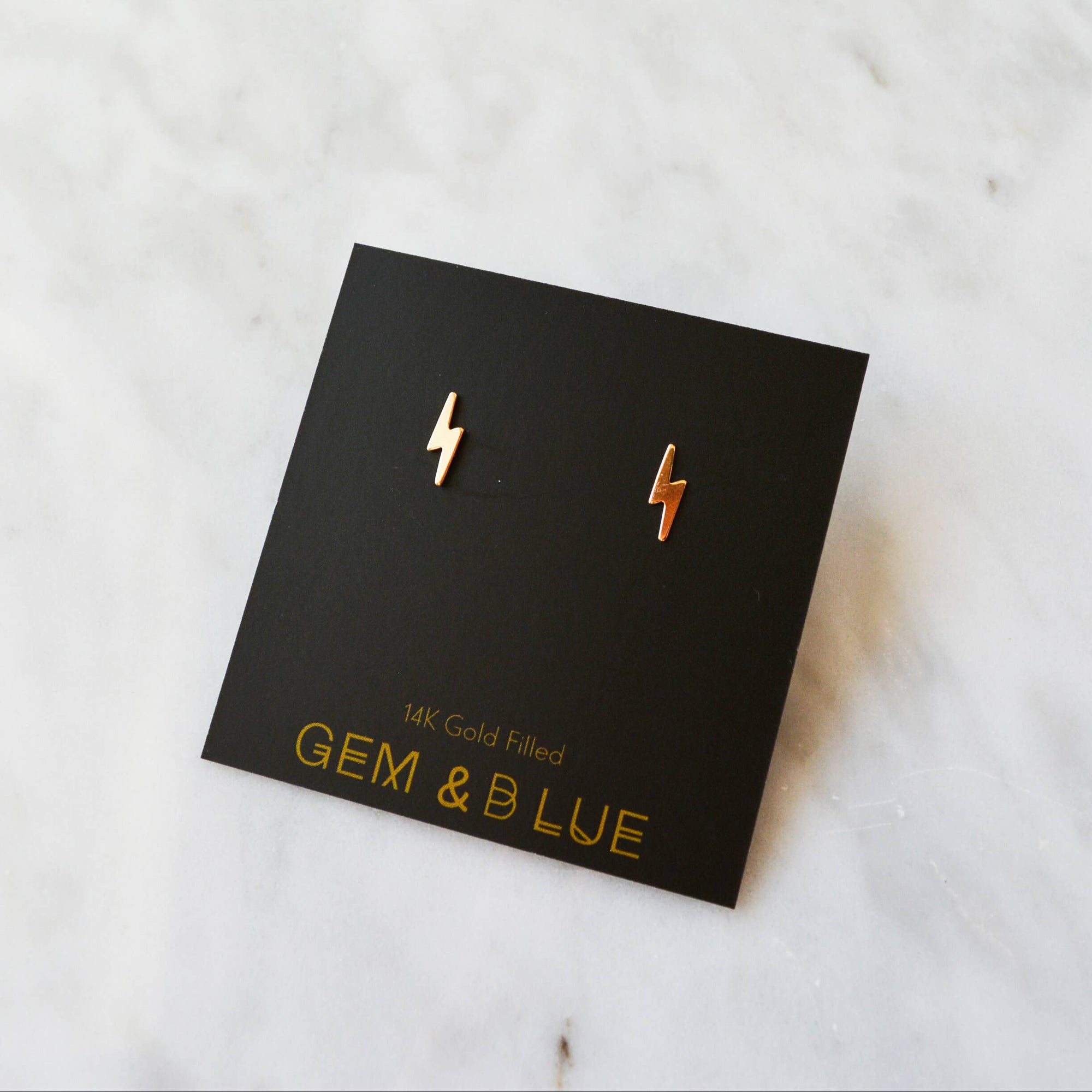 Gold lightning bolt earrings on a black card with 'GEM & LUE' branding on a marble surface.