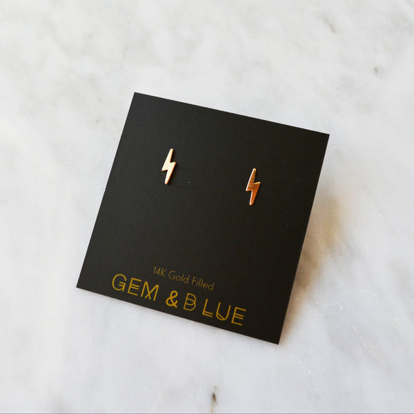Gold lightning bolt earrings on a black card with 'GEM & LUE' branding on a marble surface.