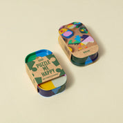 Two small round tins of Block Puzzle Me Happy 60 piece wooden jigsaw puzzle on a light beige background. One tin is open, showing colorful wooden puzzle pieces inside, and the other is closed with a kraft paper sleeve printed with a jigsaw puzzle graphic.