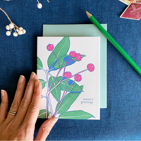 Greeting card with floral design on a blue surface with a hand, pencil, and decorative elements.