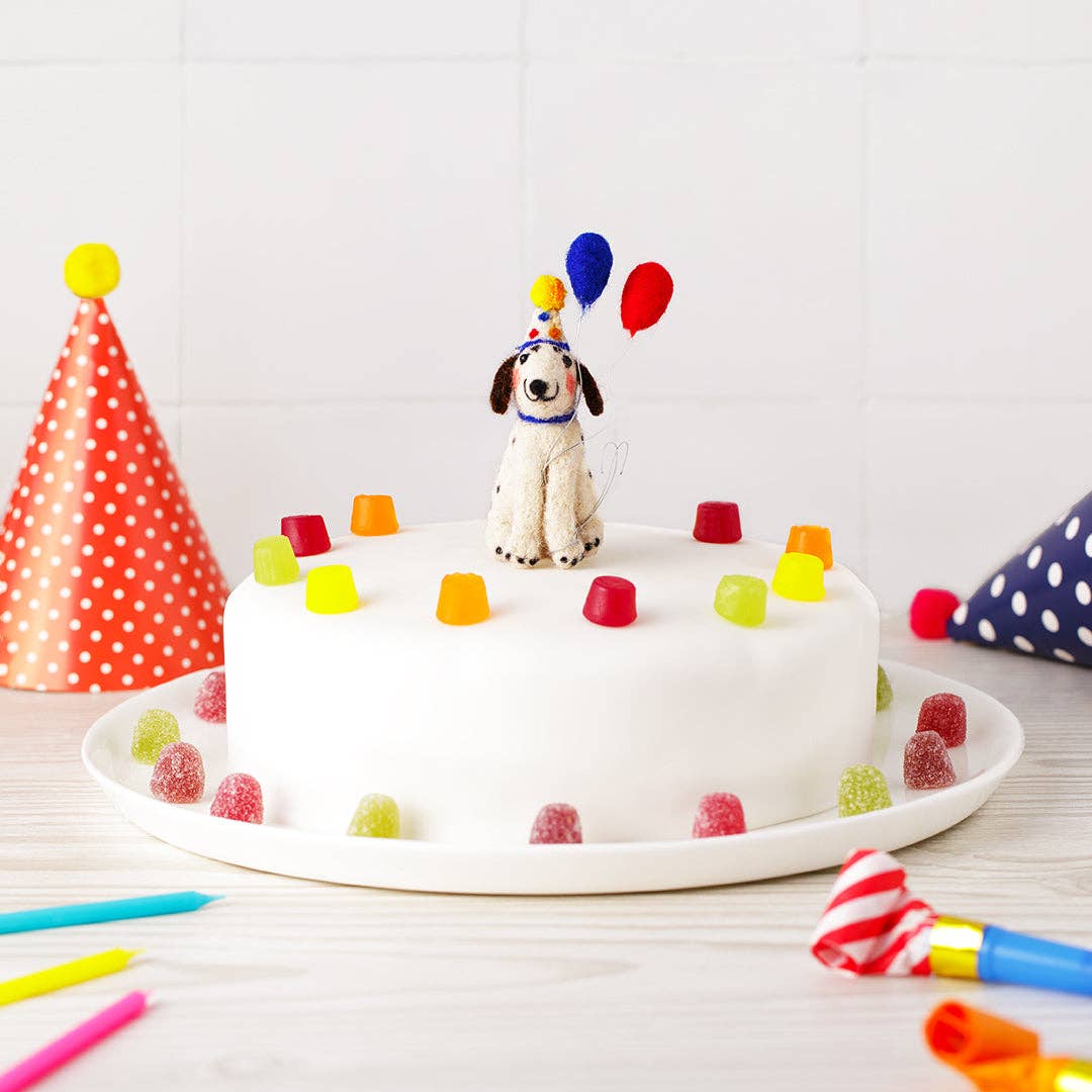 White birthday cake with dog topper and colorful decorations on a white plate, surrounded by party hats and candles.