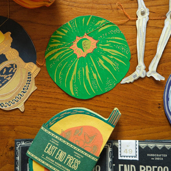 A styled flat-lay of handcrafted end press paper products on a wooden tabletop, including a round green and orange coaster, a stack of orange and yellow paper sheets wrapped with a green paper band reading 'EAST END PRESS', and other decorative paper pieces and skeleton props.