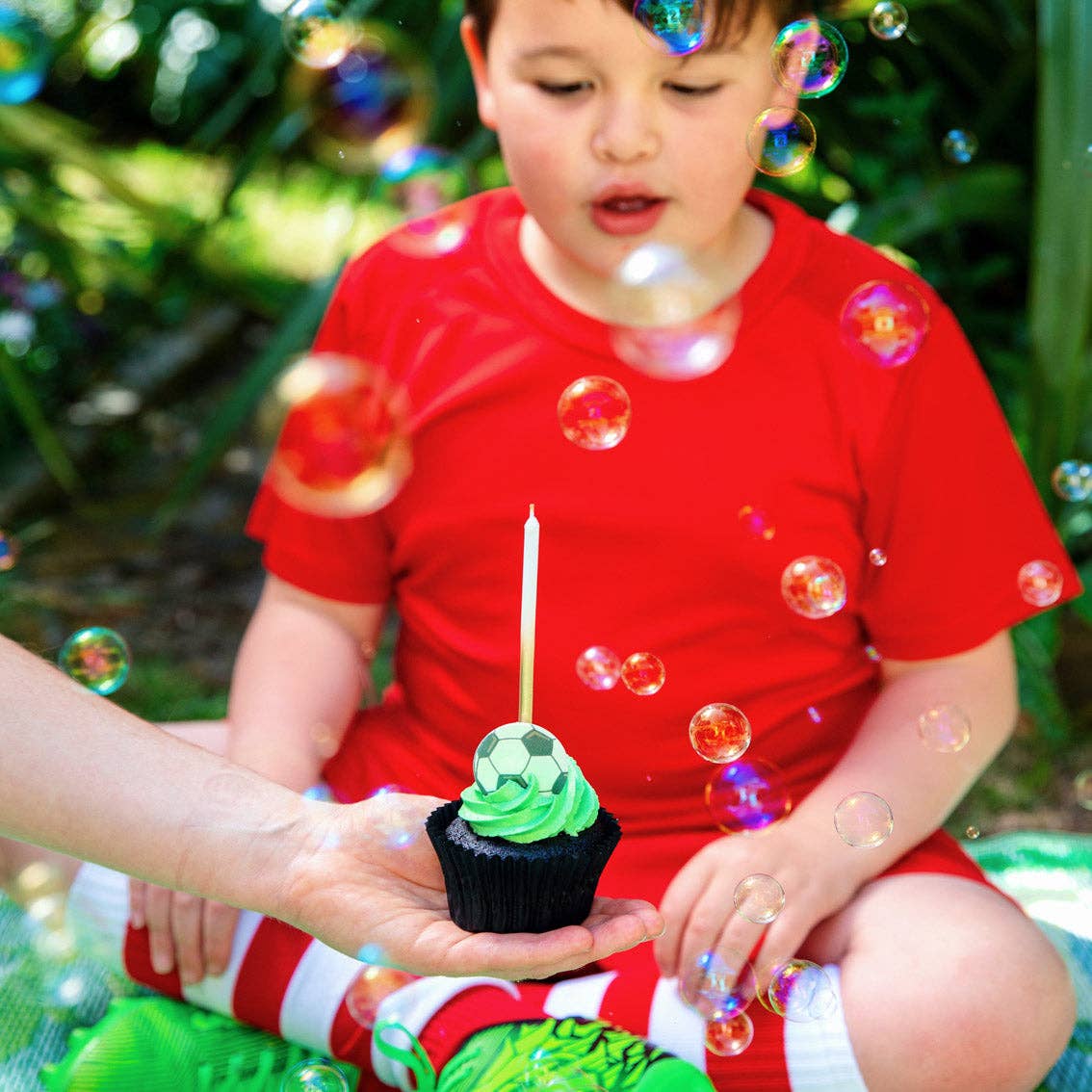 Child in a red shirt holding a cupcake with a candle, surrounded by bubbles outdoors.
