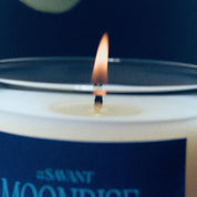 A close-up of a lit candle with a white wax surface, a single wick, and a dark navy label reading "#SAVANT MOONDIVE" against a dark navy background.