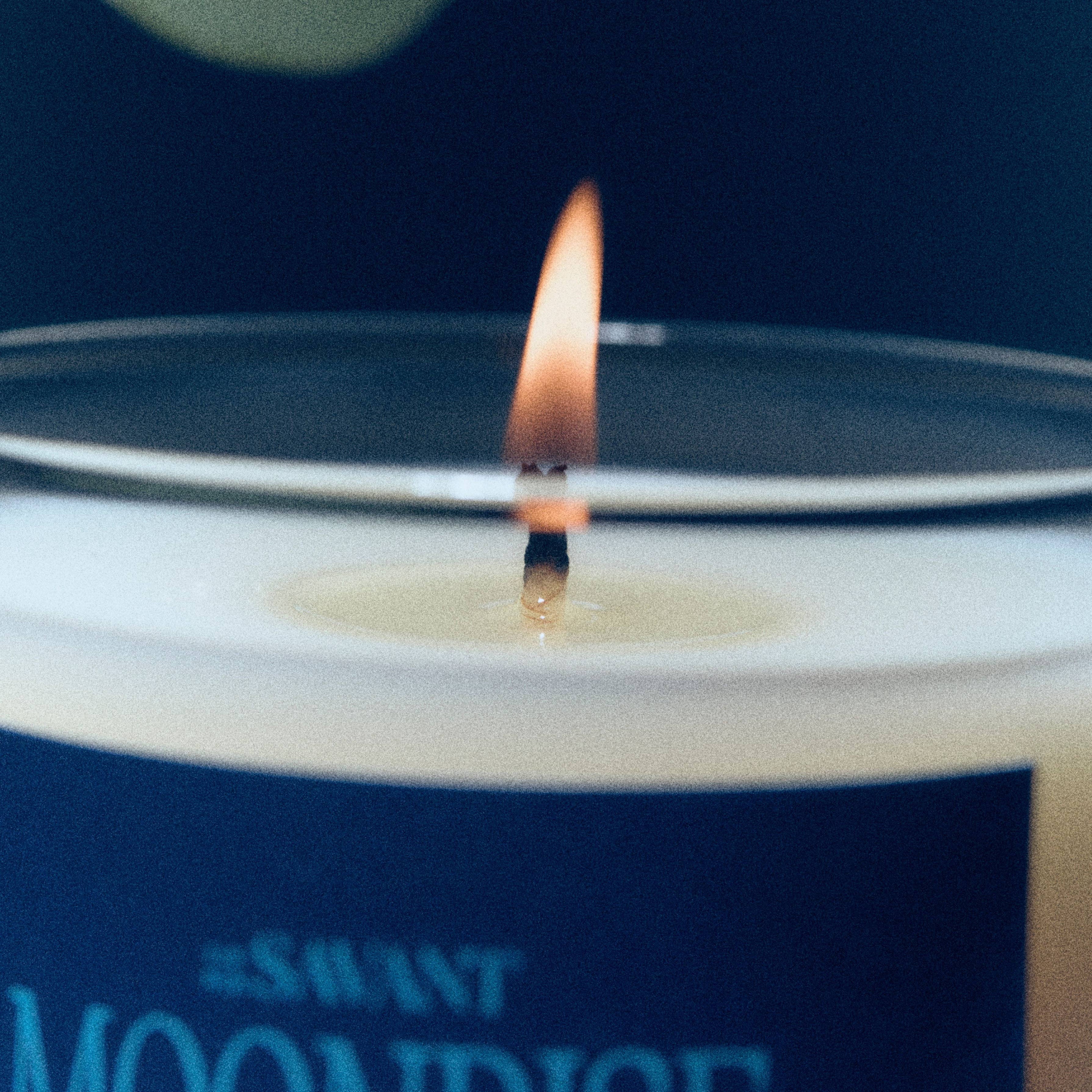 A close-up of a lit candle with a white wax surface, a single wick, and a dark navy label reading "#SAVANT MOONDIVE" against a dark navy background.