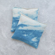 Two square fabric lavender pillows with a blue and white ocean wave pattern, displayed on a textured gray background.