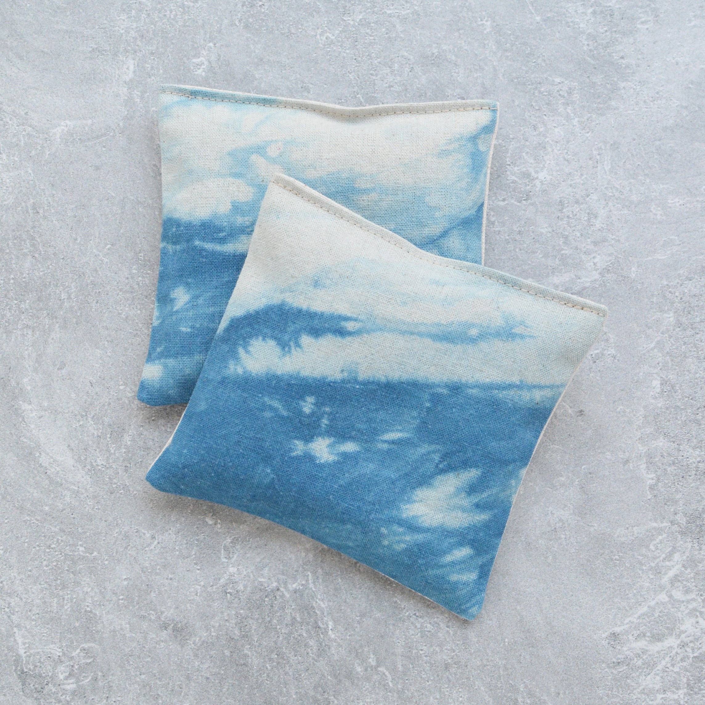 Two square fabric lavender pillows with a blue and white ocean wave pattern, displayed on a textured gray background.