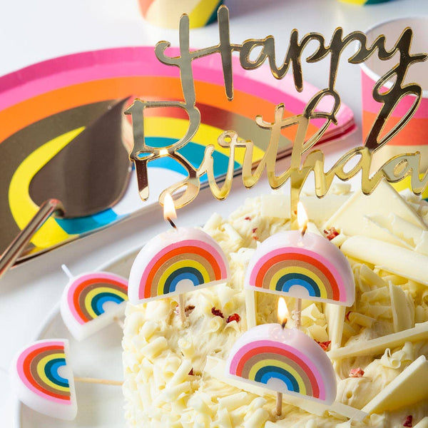 Birthday cake with rainbow candles and a 'Happy Birthday' topper on a colorful plate background.