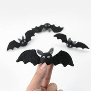 A hand holding a black felt bat finger puppet in the foreground, with several more black felt bat finger puppets arranged in the background on a plain white studio background.