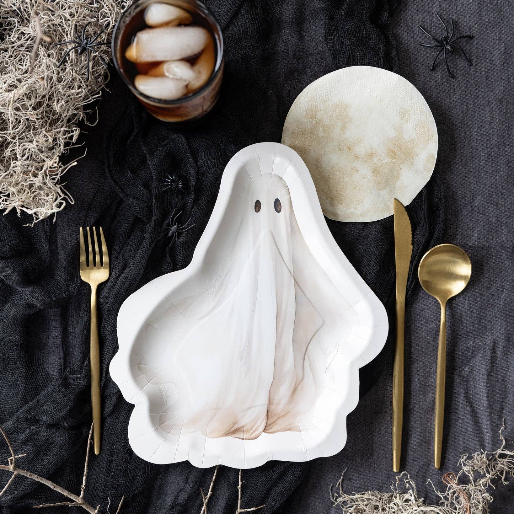 A ghost-shaped white ceramic plate with a subtle marbled texture, styled in a flat-lay with gold-toned cutlery, a glass of iced coffee, and Halloween-themed props on a dark fabric background.