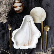 A ghost-shaped white ceramic plate with a subtle marbled texture, styled in a flat-lay with gold-toned cutlery, a glass of iced coffee, and Halloween-themed props on a dark fabric background.