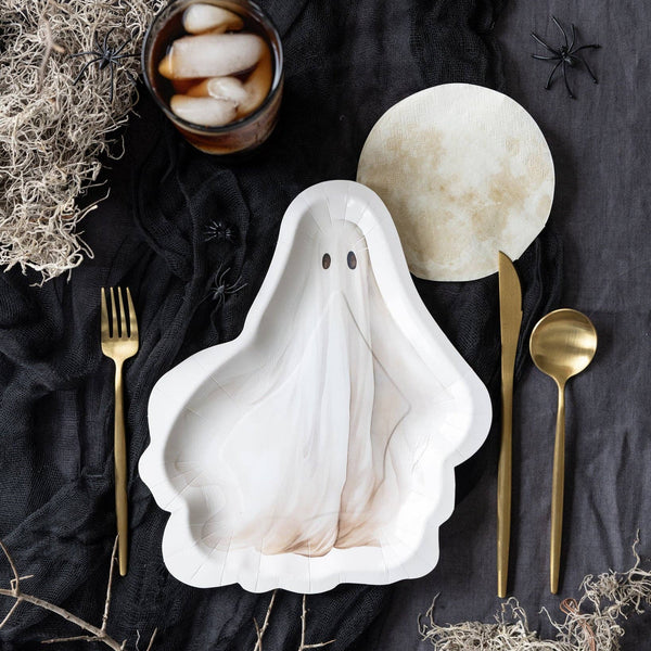 A ghost-shaped white ceramic plate with a subtle marbled texture, styled in a flat-lay with gold-toned cutlery, a glass of iced coffee, and Halloween-themed props on a dark fabric background.
