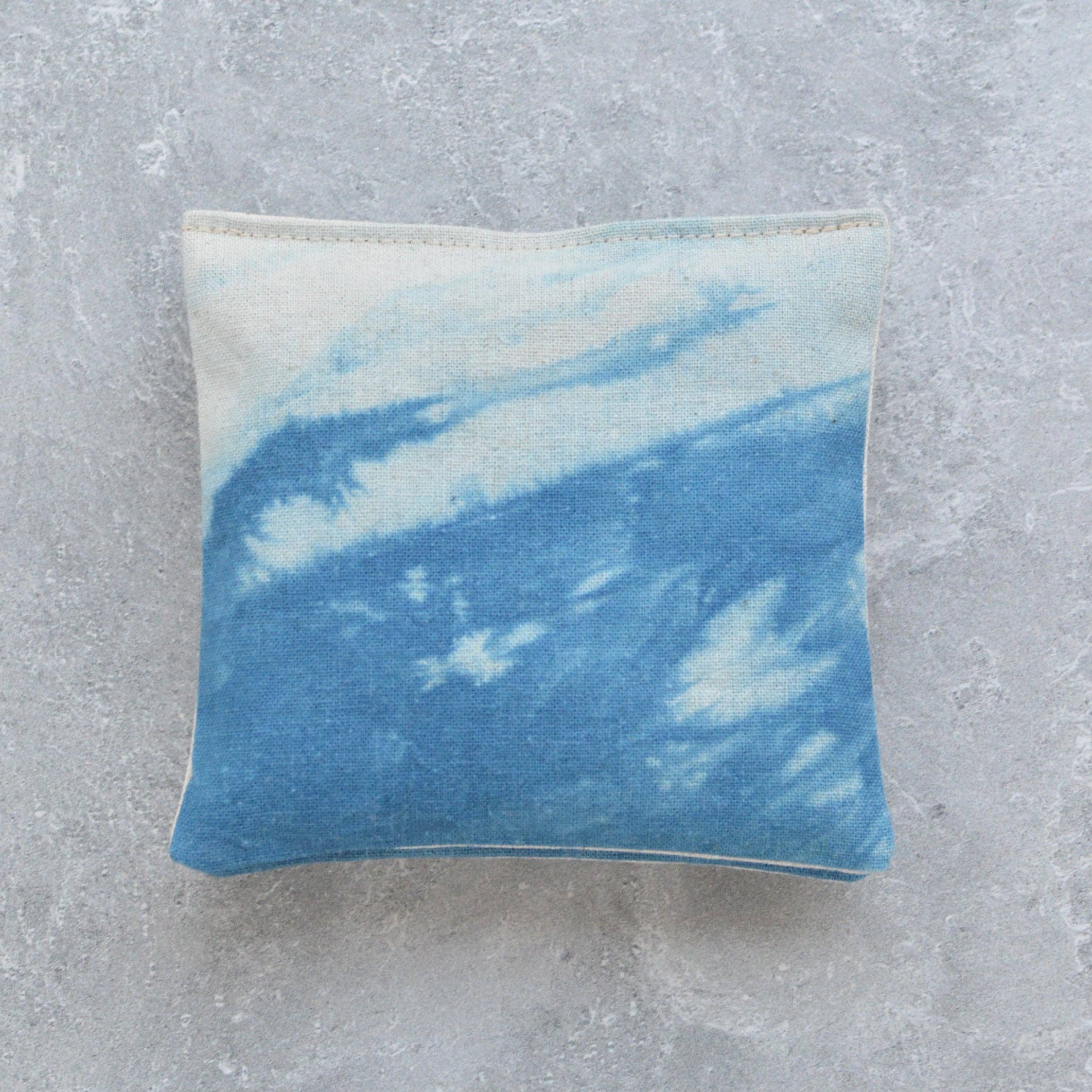 A square lavender sachet featuring a blue and white tie-dye or watercolor pattern, displayed on a textured gray concrete surface.