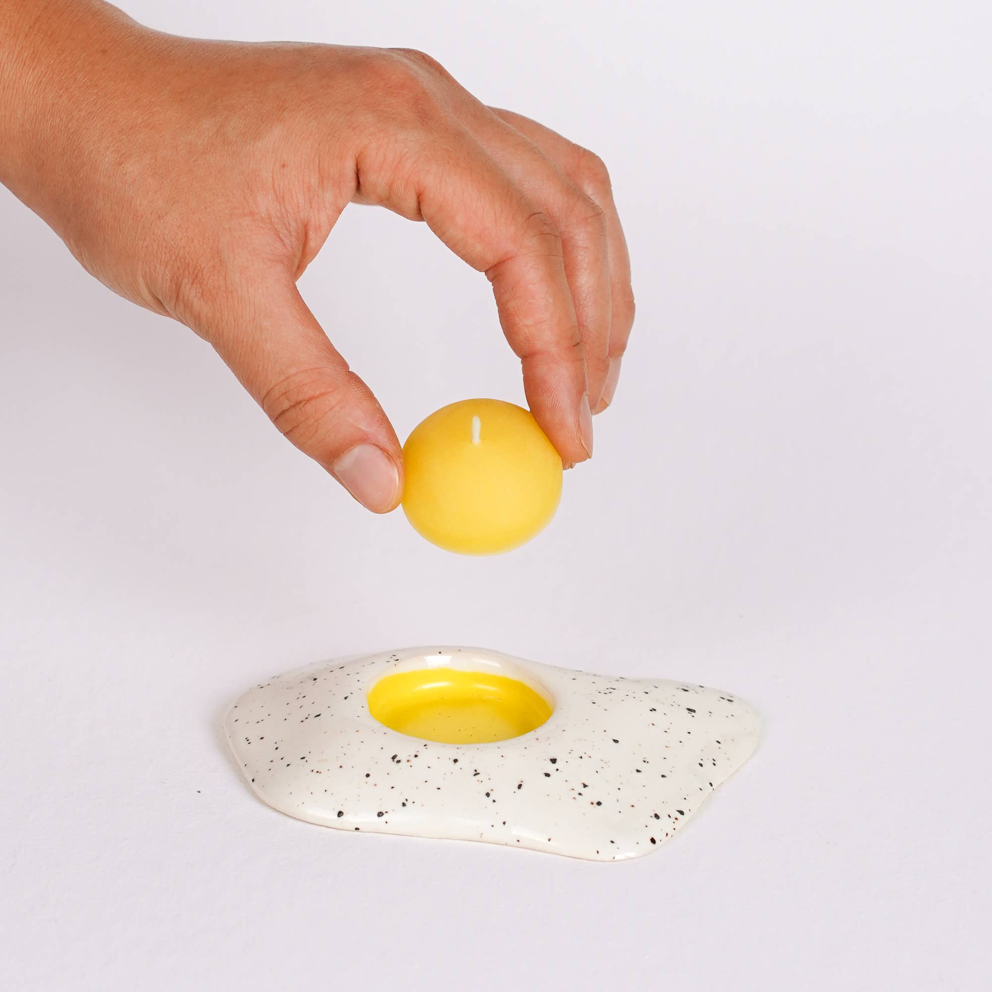 Hand placing a yellow candle on an egg candle with black speckles against a white background