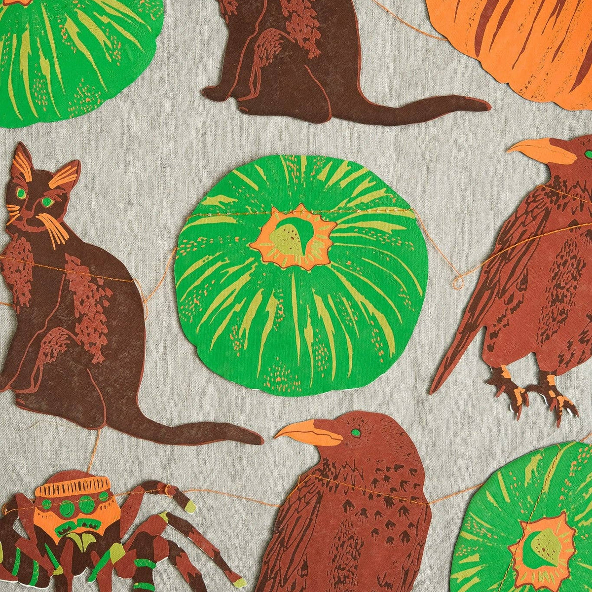 A close-up of a fabric or wallpaper pattern featuring Halloween-themed illustrations, including brown cats, orange pumpkins, green pumpkins, a brown bird, a spider, and a witch hat, all printed on a light gray textured background.