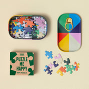 A colorful 6-piece jigsaw puzzle set by Things Books, featuring a tin-shaped puzzle box with a pull-tab lid, a small product box reading 'PUZZLE ME HAPPY', several loose puzzle pieces, and a tray showing the assembled puzzle design, all arranged on a warm beige background.