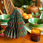 A small decorative Christmas tree made from layered patterned paper stands on a wooden dining table, surrounded by green bowls, amber glasses, napkins, and holiday decor in a warm indoor setting.
