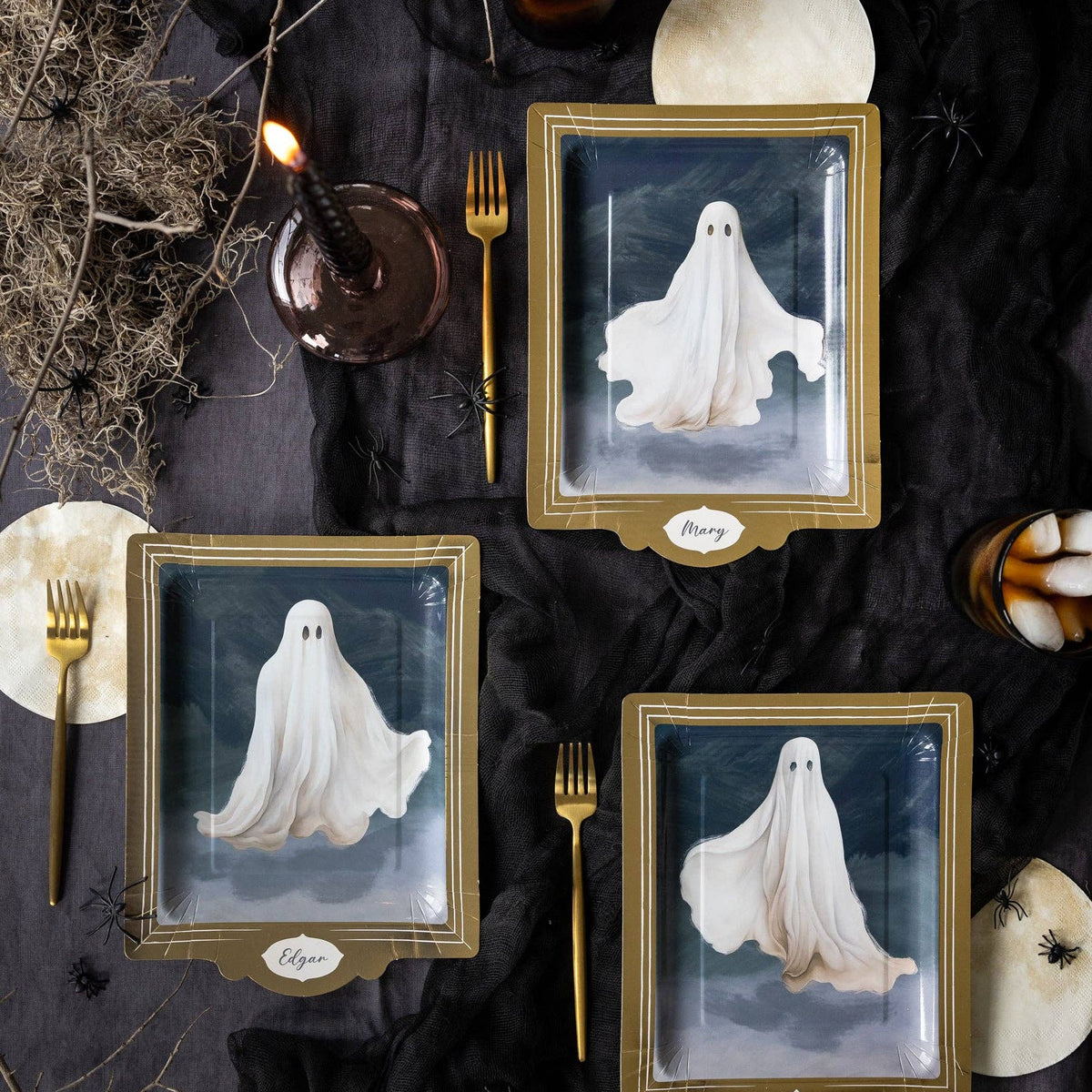 Three rectangular gold-framed plates with clear lids, each showing a white ghost illustration, arranged on a black fabric table setting with gold forks, candles, dried branches, and moon decorations.