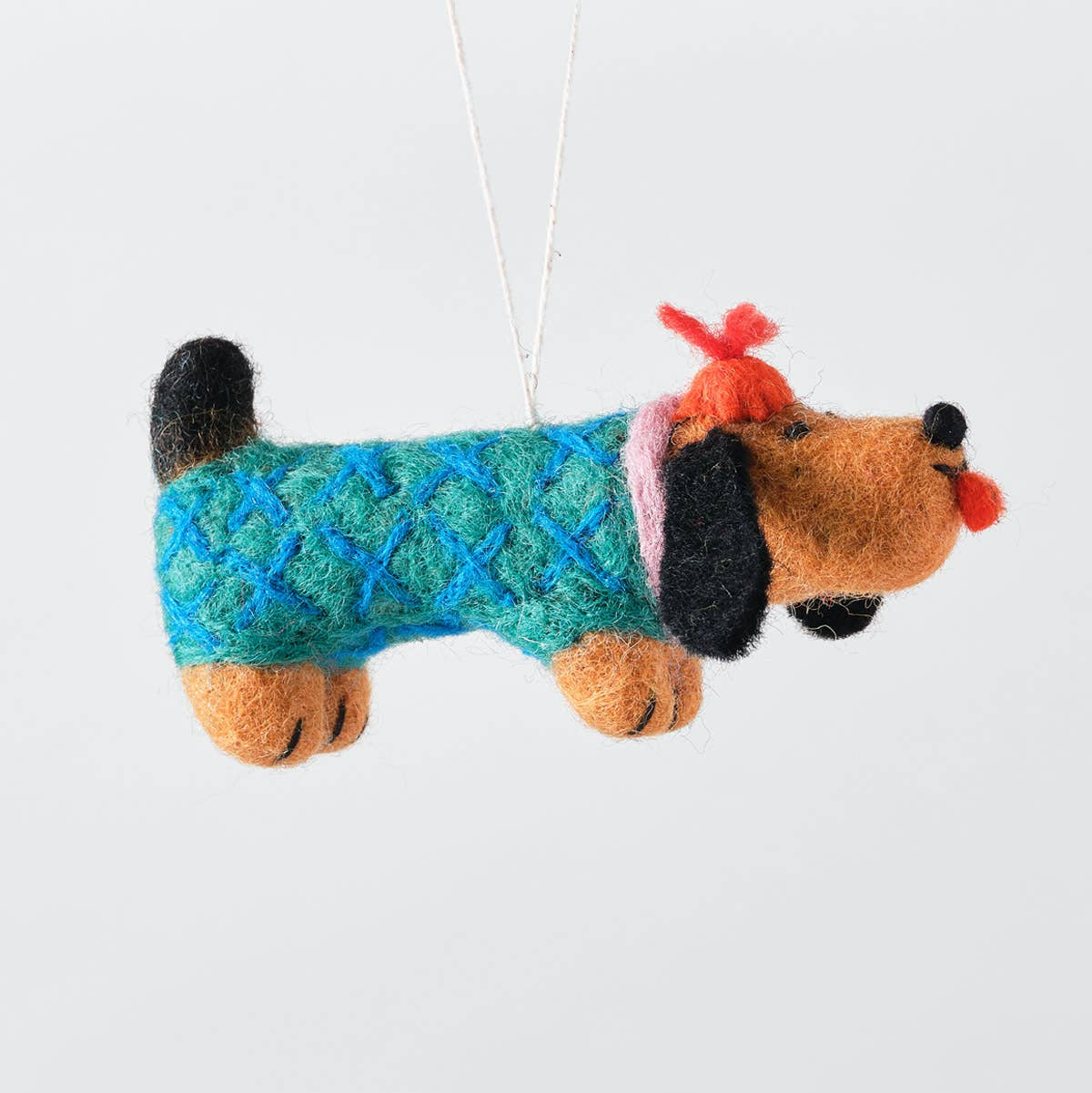 A felt dog ornament wearing a blue and green patterned sweater, with black ears and tail, orange paws and snout, and a red bow on a white background.