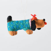 A felt dog ornament wearing a blue and green patterned sweater, with black ears and tail, orange paws and snout, and a red bow on a white background.