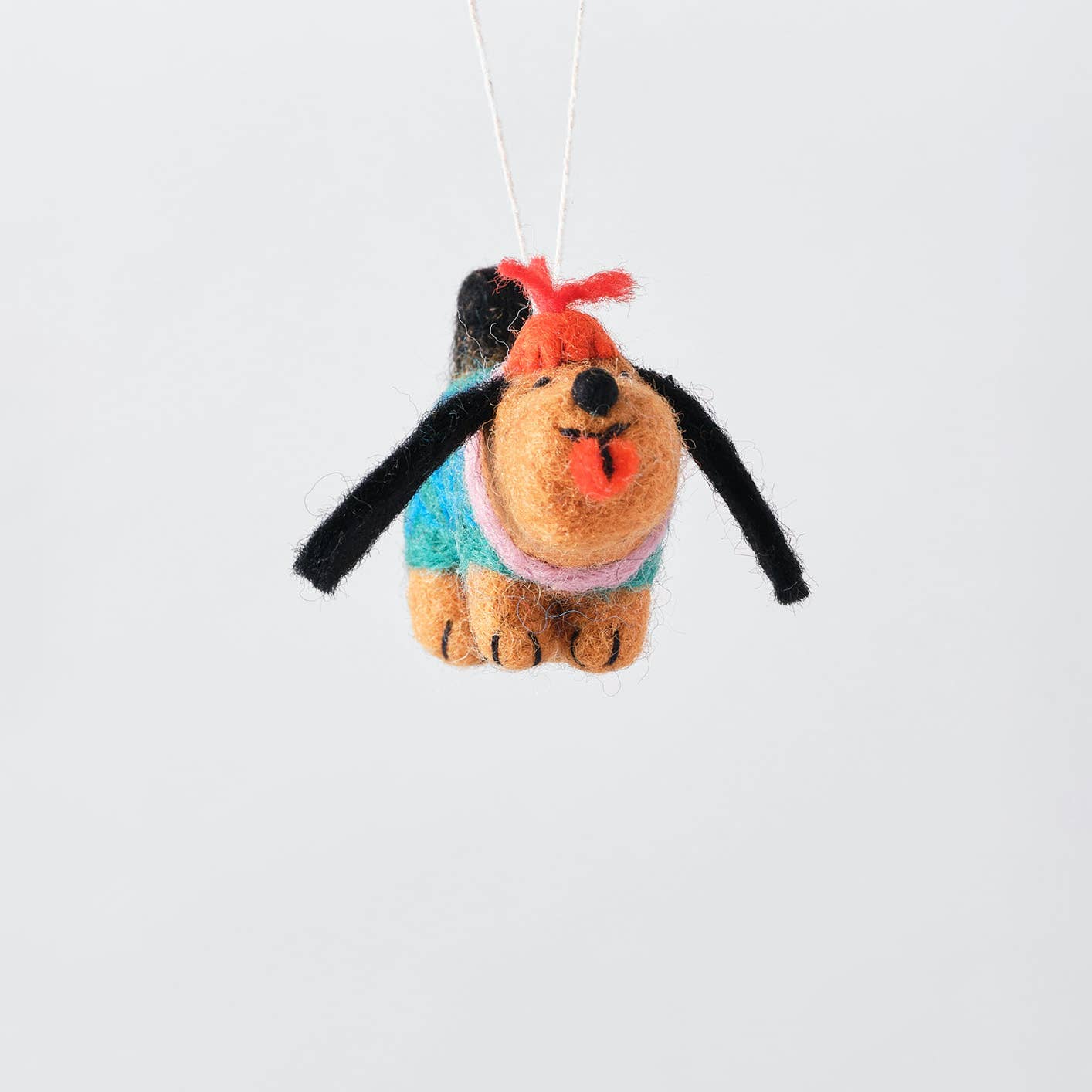 A handcrafted felt dog ornament hanging from a white string against a clean, light gray background. The dog features tan fur, black floppy ears, a turquoise sweater, a pink collar, and a red bow on its head.