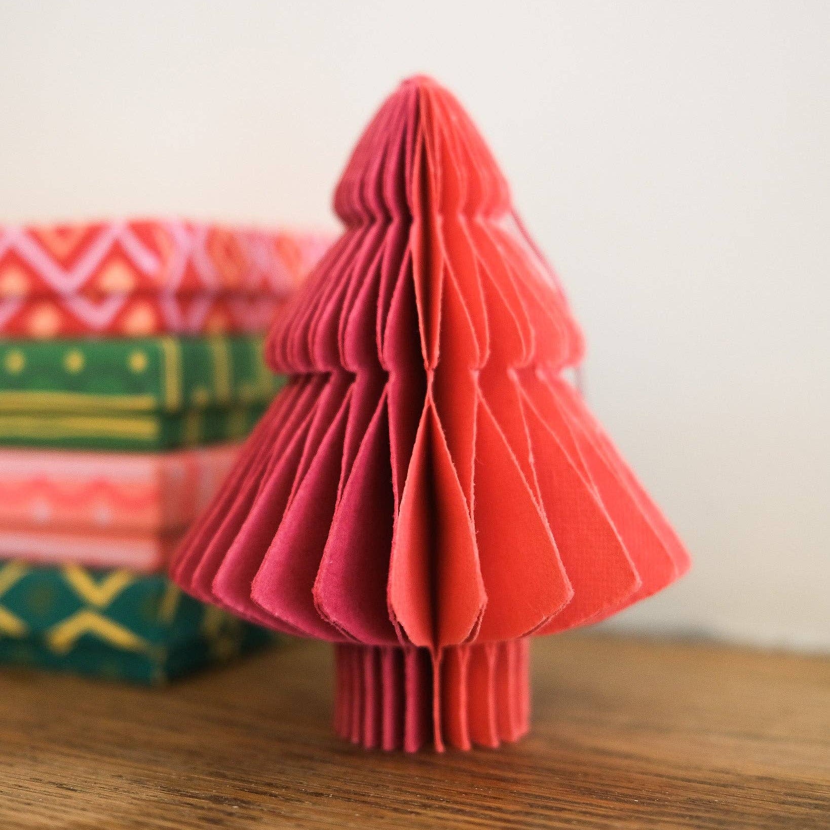 Red paper Christmas tree on a wooden surface with colorful gift boxes in the background.