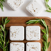 Square white soap bars with herbs on a wooden tray, surrounded by rosemary leaves.