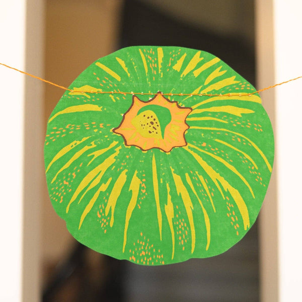 A circular green and yellow decorative paper cutout hanging on a string in front of a blurred indoor doorway background.