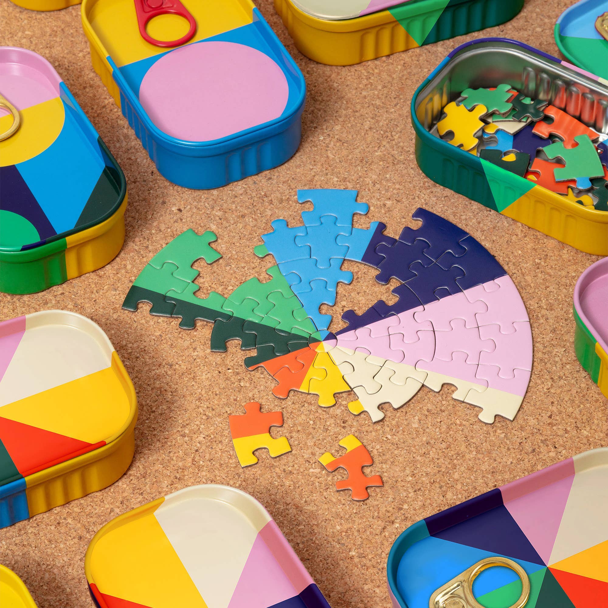 A styled flat-lay of colorful jigsaw puzzle pieces arranged in a partial circular pattern on a cork surface, surrounded by matching pastel-colored metal tin containers.