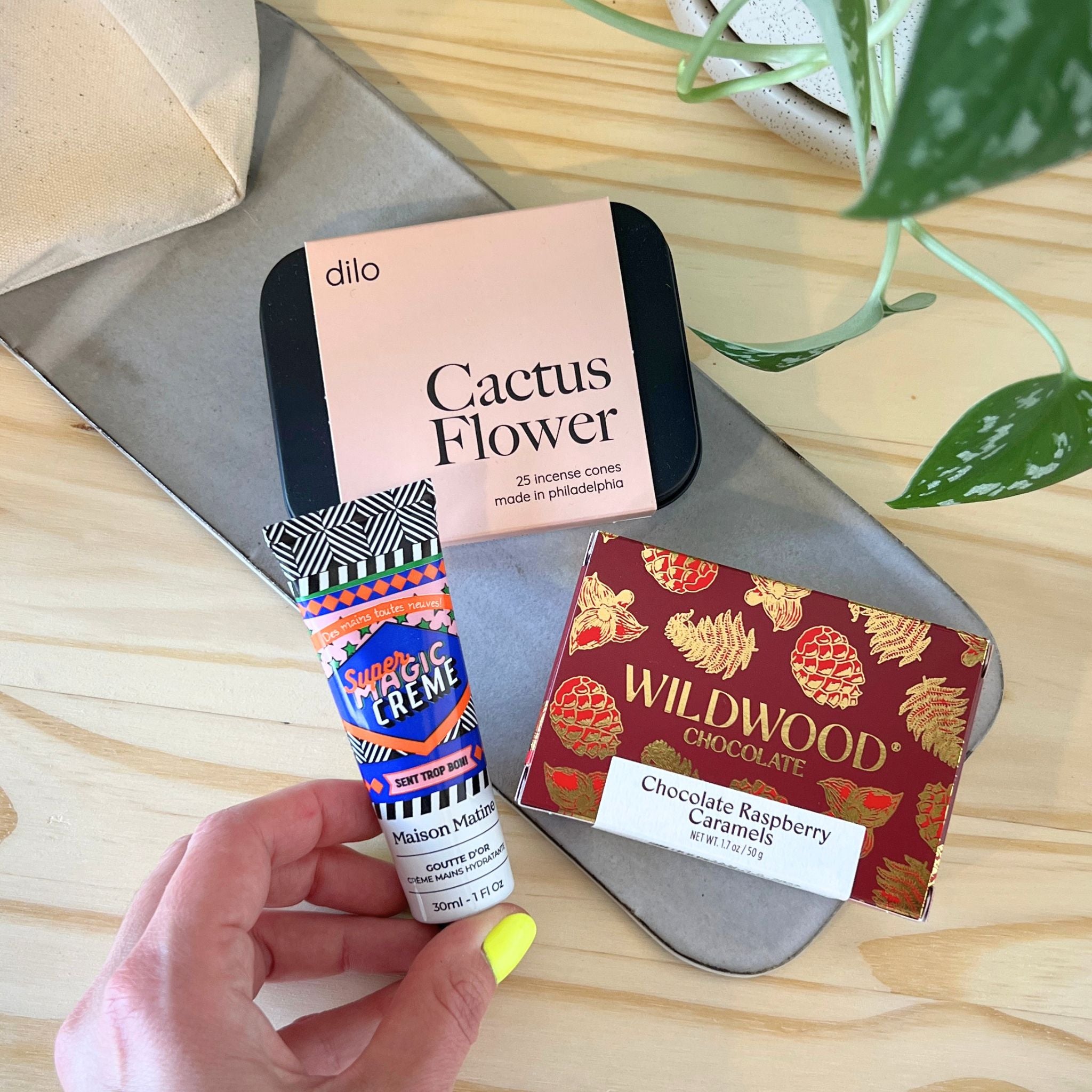 Hand holding a tube of Hand Creme with a box of Dillo Cactus Flower and Wildwood Chocolate on a wooden surface.
