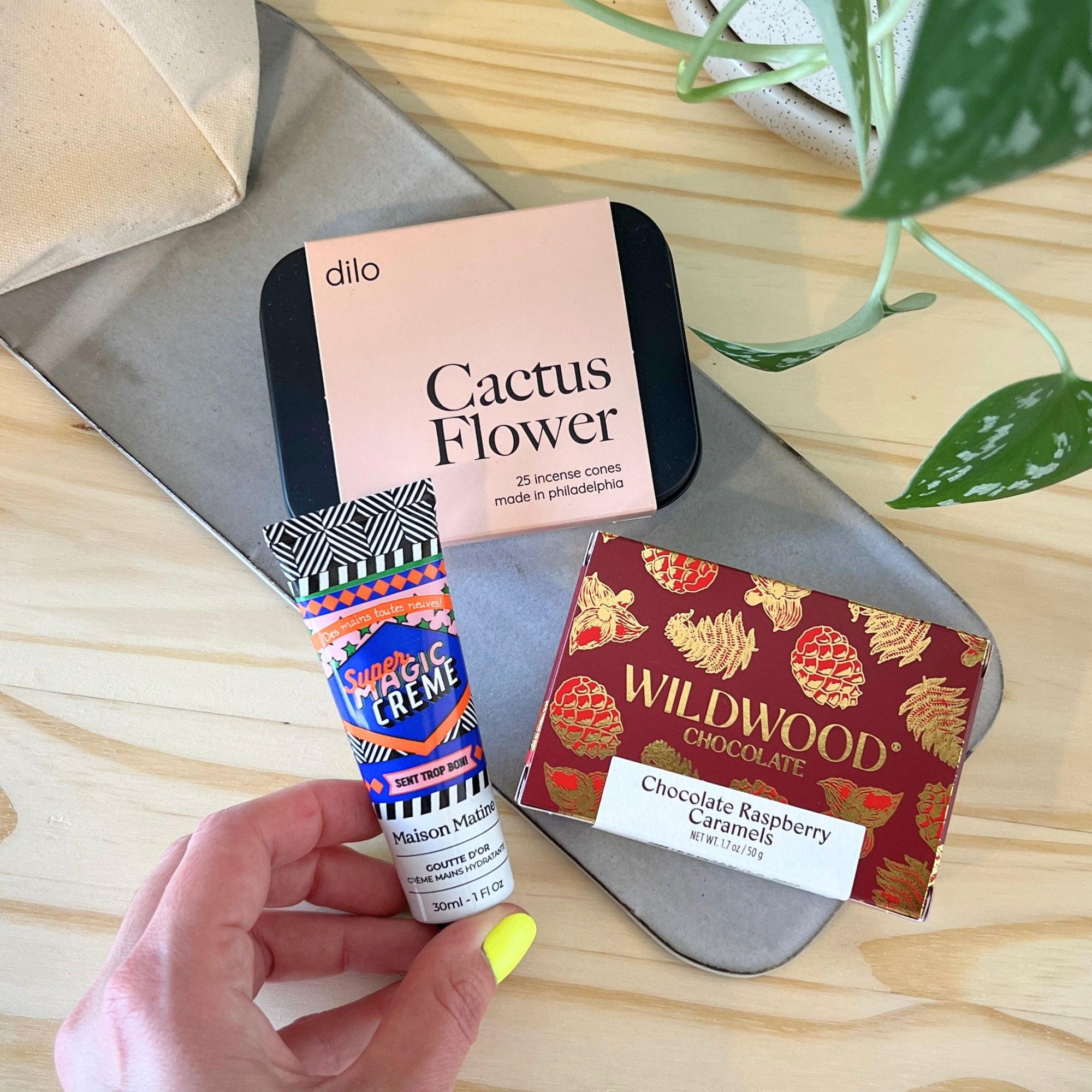 Hand holding a tube of Hand Creme with a box of Dillo Cactus Flower and Wildwood Chocolate on a wooden surface.