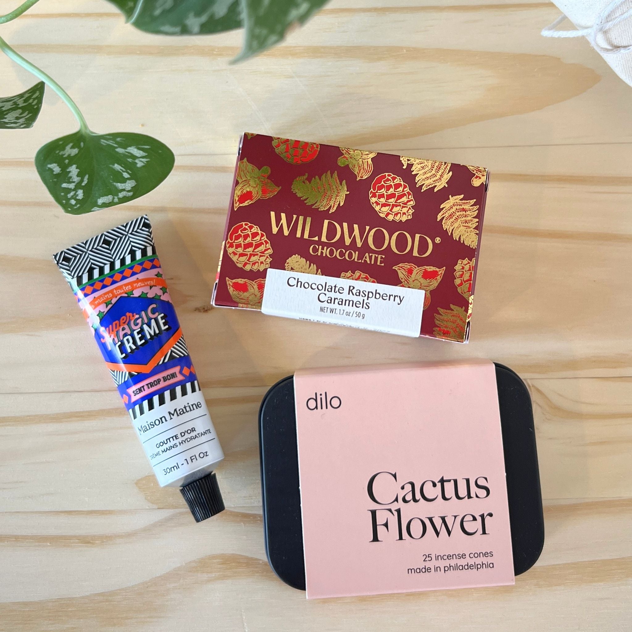 Three products on a wooden surface: a tube of Magic Creme, a box of Wildwood Chocolate, and a container of Dilo Cactus Flower incense cones.