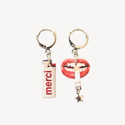 Two dangly earrings, one featuring a white lighter shape with the word "MERCI" written on it, the other earring a pair of lips with a cigarette hanging from it, a star at the end of the cigarette. Against a light grey background.