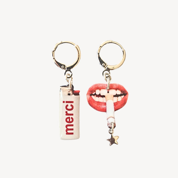Two dangly earrings, one featuring a white lighter shape with the word "MERCI" written on it, the other earring a pair of lips with a cigarette hanging from it, a star at the end of the cigarette. Against a light grey background.