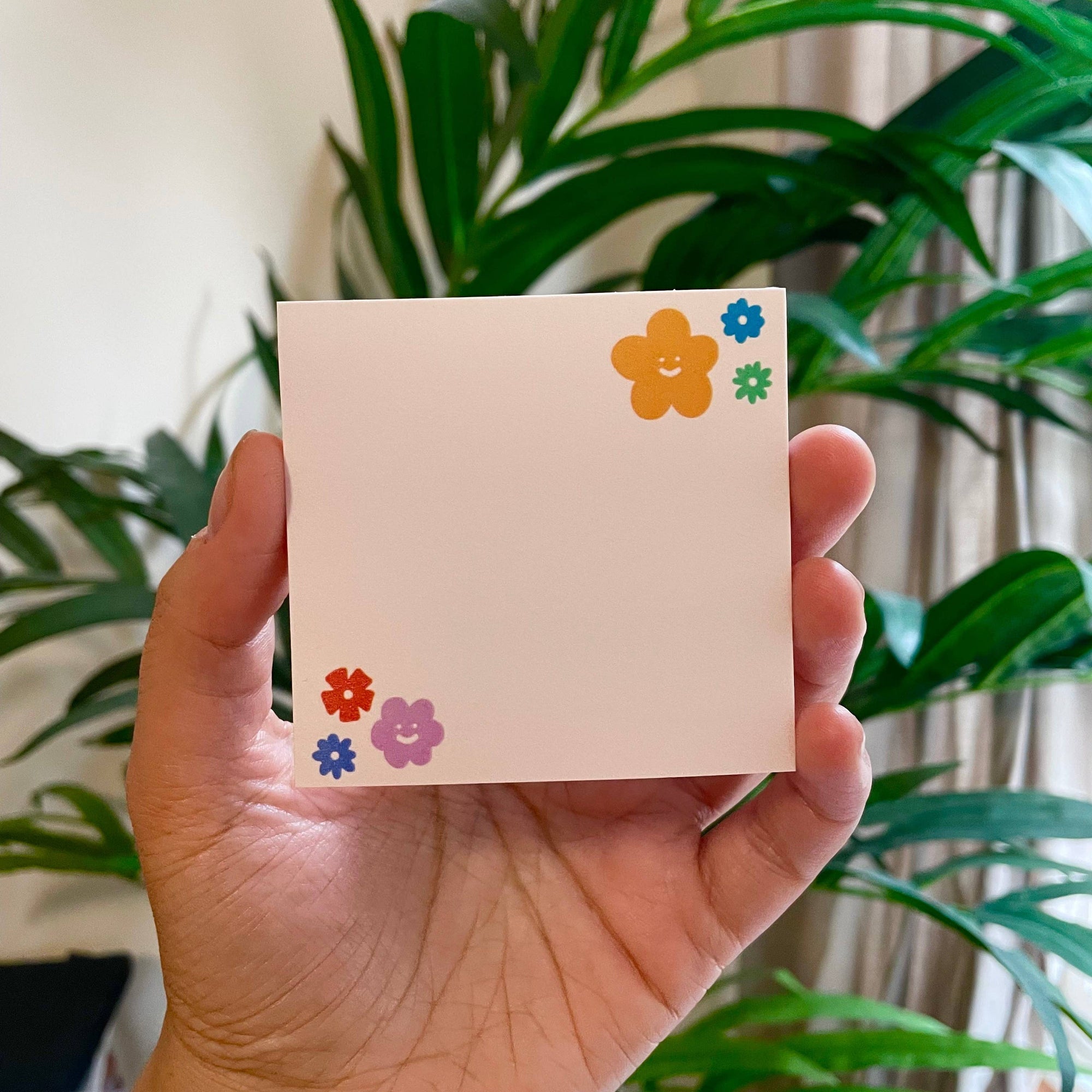 Hand holding a small square card with colorful flower designs against a green plant background