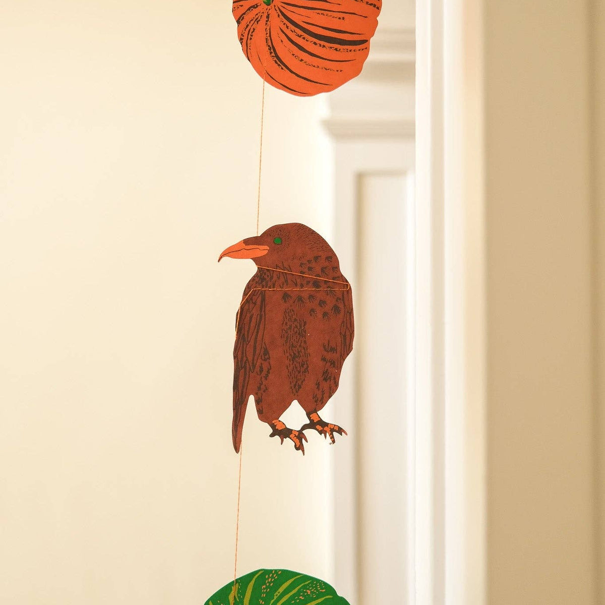 A hanging autumn decoration featuring three illustrated paper-cut style shapes: an orange pumpkin at the top, a brown bird in the middle, and a green pumpkin at the bottom, suspended by thin strings against a warm beige interior wall.