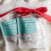 Two clear pouches of Ritual No. 8 Winter Sanctuary bath tea tied with a red ribbon, placed on a textured cream knit blanket.