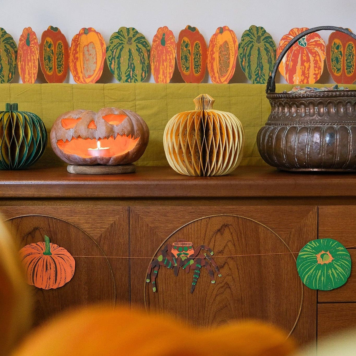 A styled autumn scene featuring decorative paper pumpkins and a hanging basket arranged on a wooden sideboard, with a blurred orange foreground in the foreground.