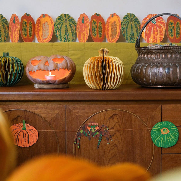 A styled autumn scene featuring decorative paper pumpkins and a hanging basket arranged on a wooden sideboard, with a blurred orange foreground in the foreground.