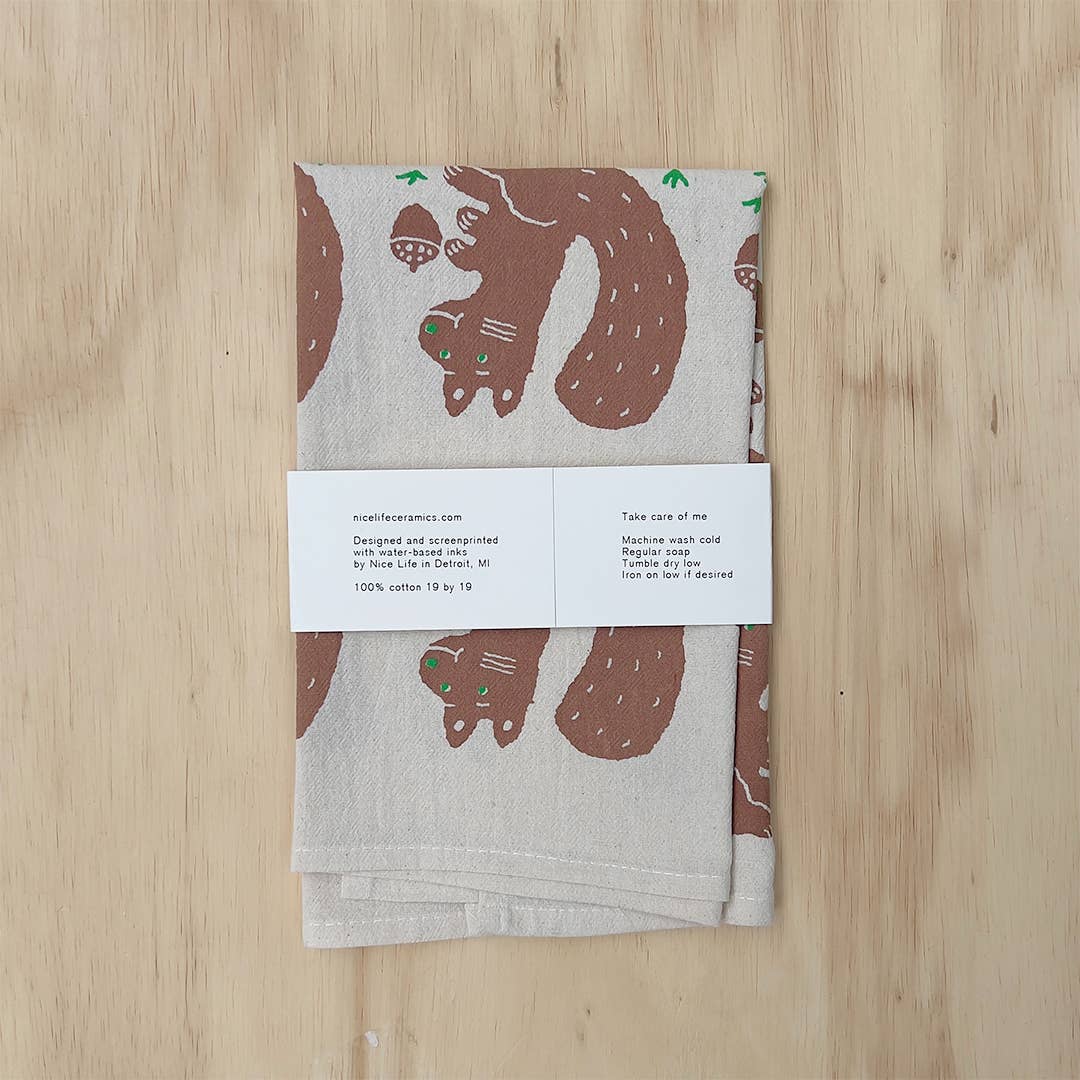 A folded cotton tea towel with a brown squirrel and acorn pattern, wrapped in a white paper band, displayed flat on a light wood background.