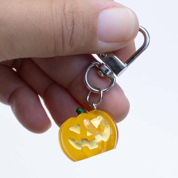 A hand holding a small yellow jack-o'-lantern keychain charm with a silver metal clasp against a plain white background.
