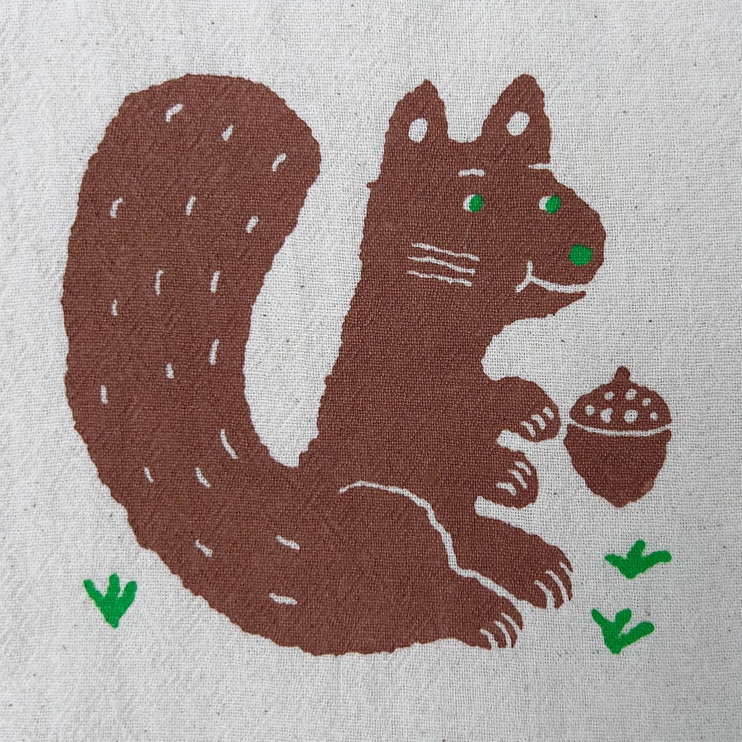 A close-up of a textured fabric featuring a stylized brown squirrel holding an acorn, with small green grass accents and green eyes.