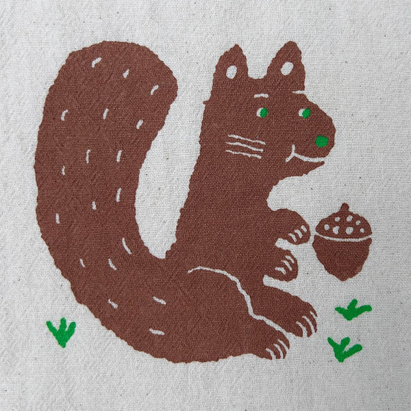A close-up of a textured fabric featuring a stylized brown squirrel holding an acorn, with small green grass accents and green eyes.