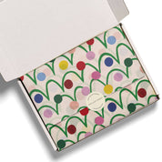 An open white cardboard gift box showing a folded envelope-style package wrapped in cream paper with a colorful abstract pattern of green lines and multicolored dots. A round white sticker on the front reads 'RED CAP CLOTHING'.