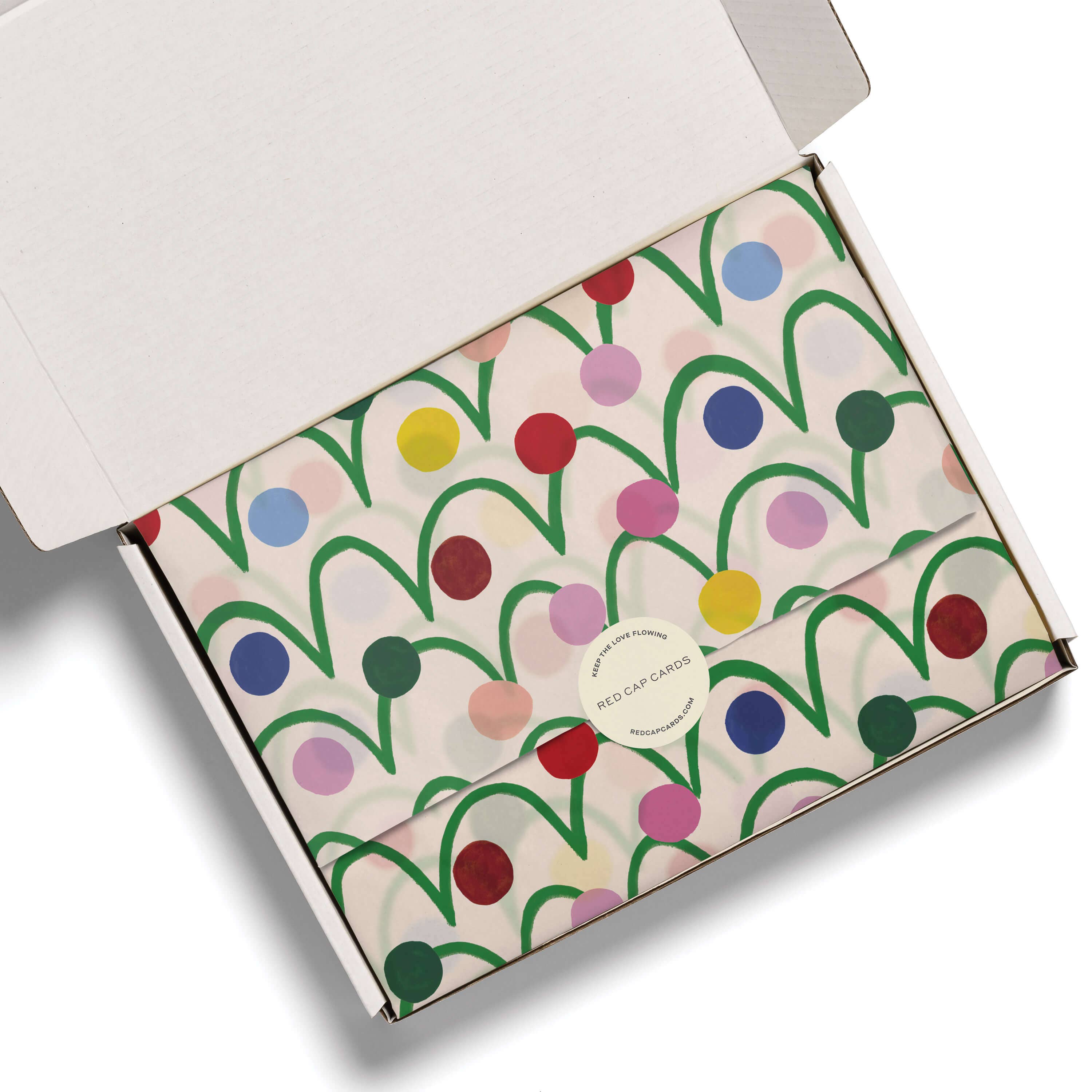 An open white cardboard gift box showing a folded envelope-style package wrapped in cream paper with a colorful abstract pattern of green lines and multicolored dots. A round white sticker on the front reads 'RED CAP CLOTHING'.