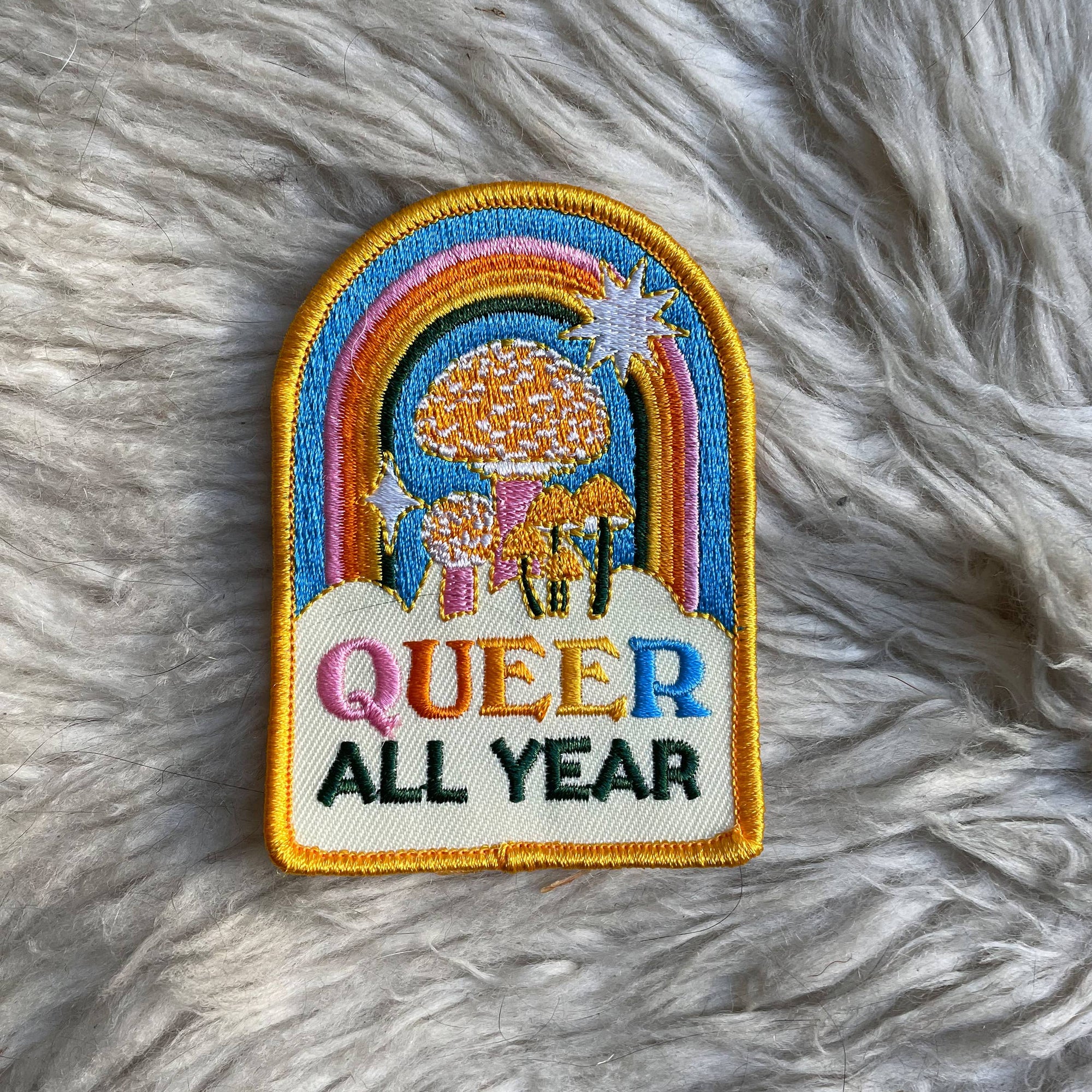 Embroidered patch with rainbow, sun, and 'Queer All Year' text on a fluffy white background