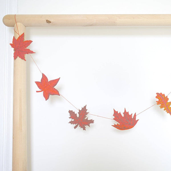 A string garland of autumn leaf cutouts in red, orange, and yellow, hanging from a light wood dowel against a plain white wall.